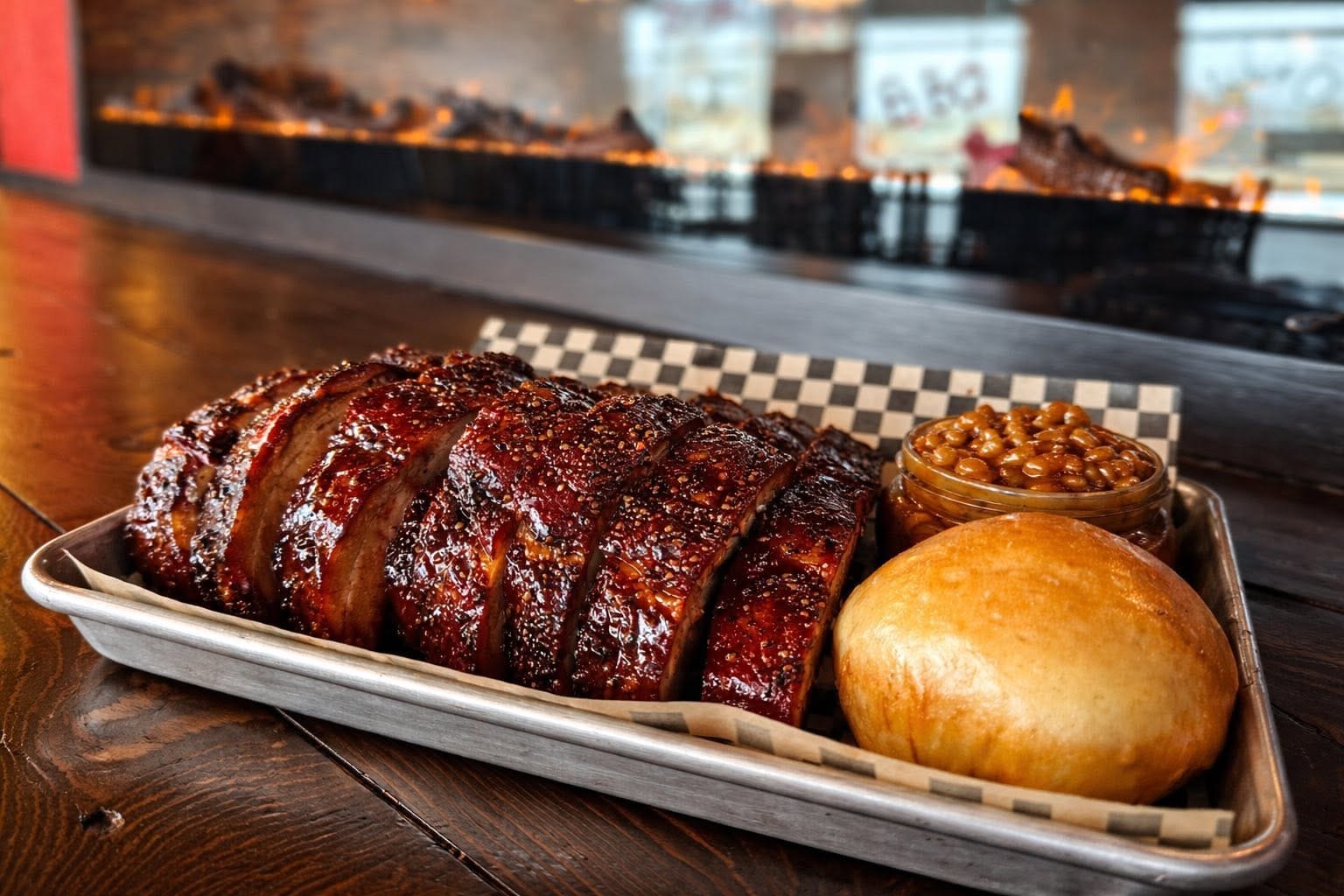Glazed sliced ribs on a tray with baked beans and a roll, with the smoker flames glowing behind